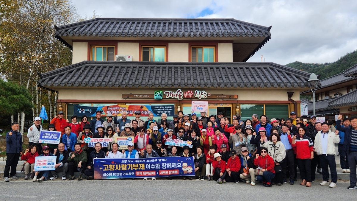 여수에서 고향 사랑 기부재 행사에 참여한 사람들로, 다양한 복장을 한 50명 이상의 사람들이 기념사진을 찍고 있다. 배경에는 전통 한옥 건물이 보인다.