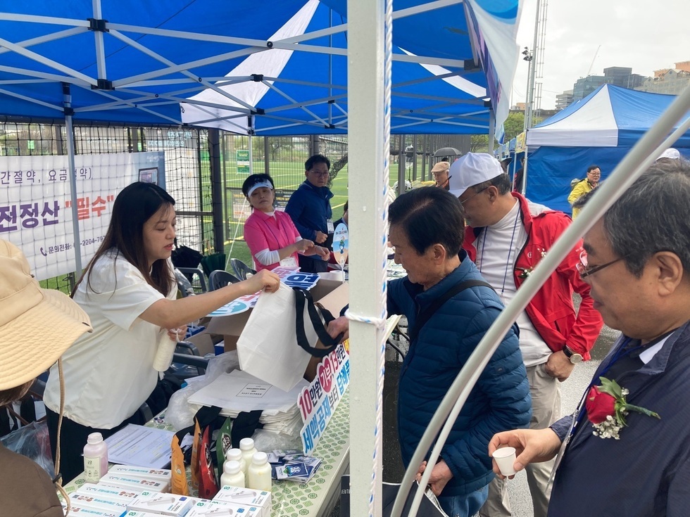 행사장에서 한 여성이 테이블에서 물품을 나누고 있으며, 남성들이 관심을 보이고 있다.