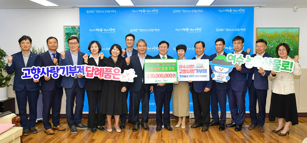 여수시청에서 고향사랑 기부제 관련 행사에 참석한 관계자들이 기념 촬영을 하고 있다. 여러 사람이 손을 들어 응원하며 기부금과 관련된 내용을 담은 현수막을 들고 있다.