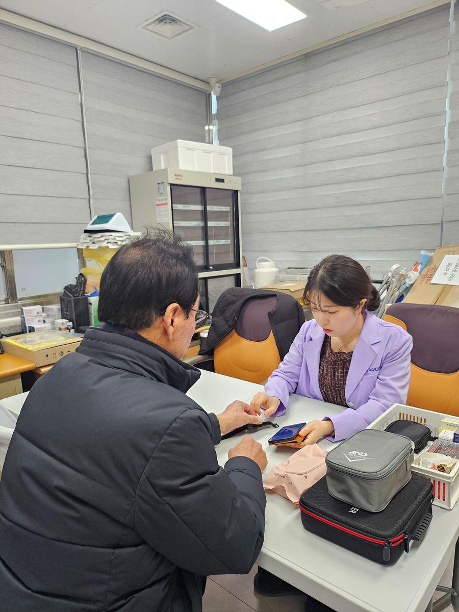 한 남성이 테이블에 앉아 있는 여성과 대화하며 스마트폰을 보여주고 있다. 여성은 보라색 의상을 입고 있으며, 주변에는 여러 가지 물품이 놓여 있다.