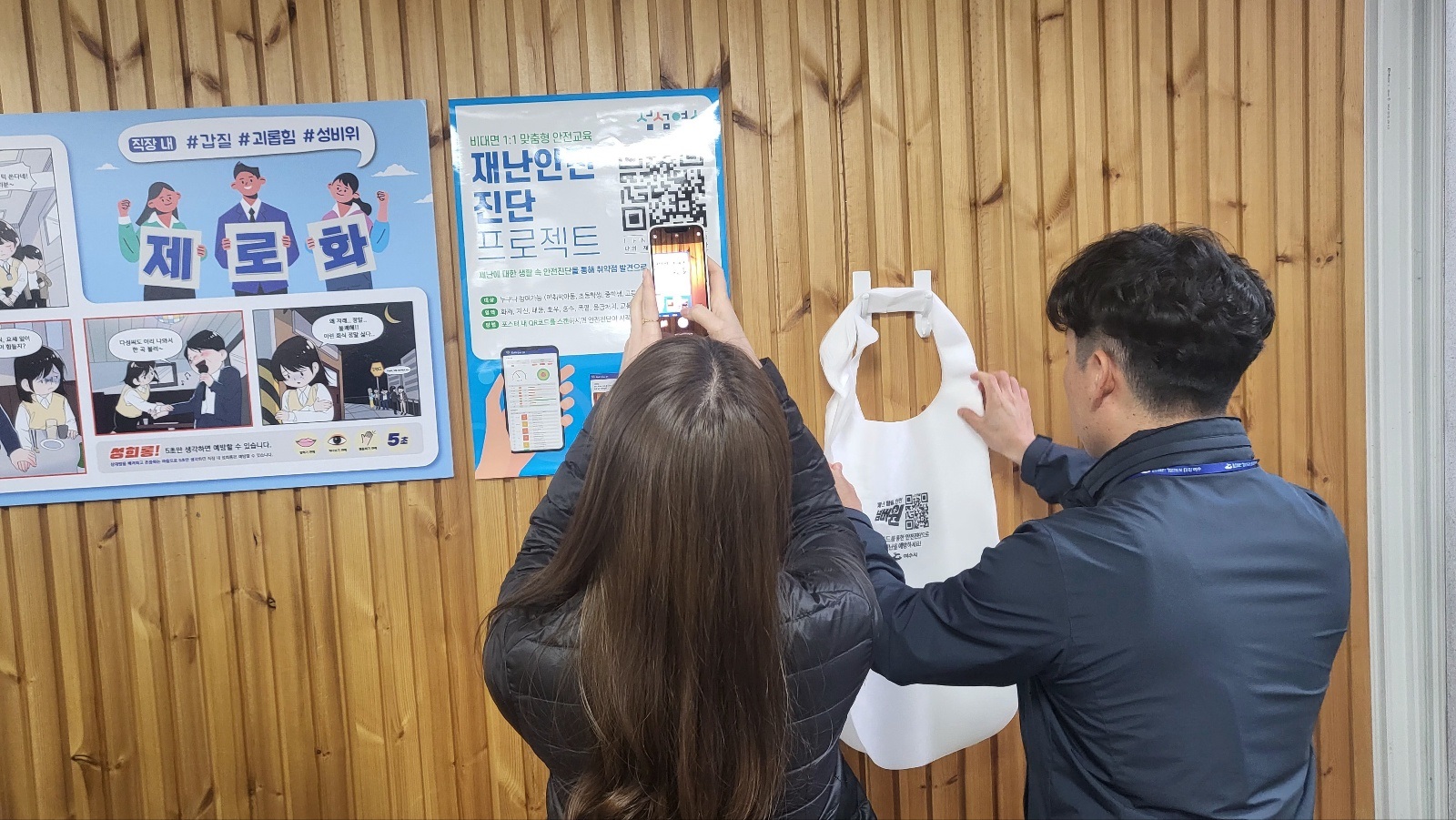 두 사람이 재난안전 진단 프로젝트와 관련된 QR코드를 스캔하고 있는 장면으로, 벽에는 관련 안내 포스터가 부착되어 있다.