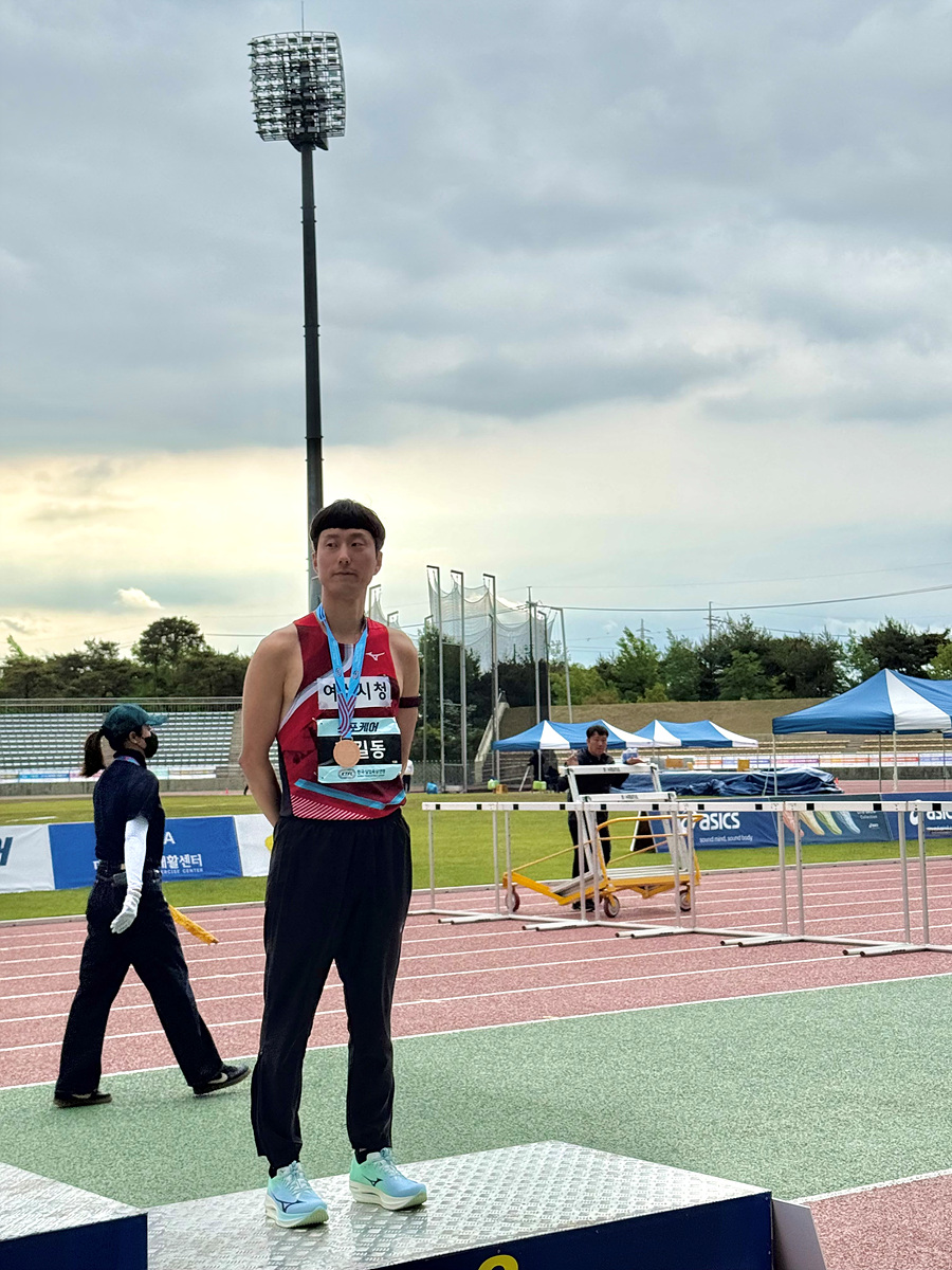 선수가 메달을 목에 걸고 시상대에 서 있는 모습으로, 배경에는 운동장이 보이고 다른 관계자들이 활동하고 있다.