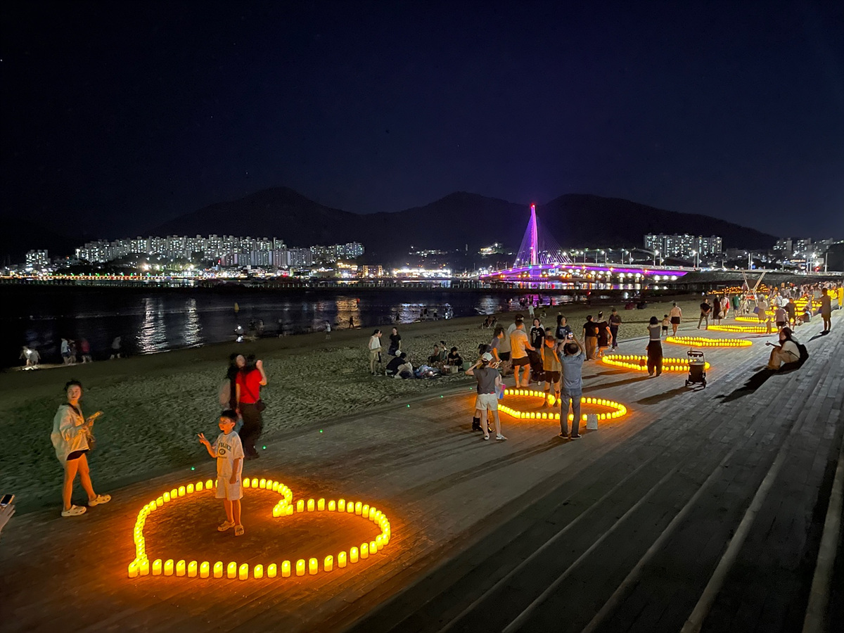 밤바다에서 사람들이 조명을 이용해 하트 모양을 만들고 있으며, 배경에는 도시의 불빛과 다리가 보인다.