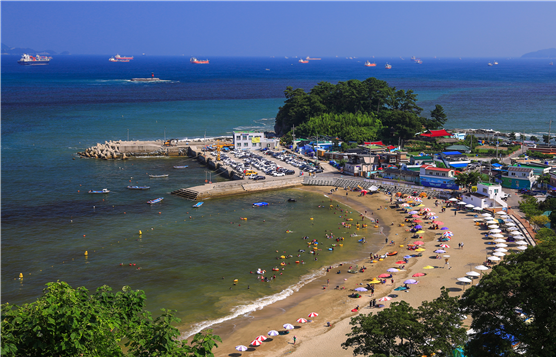 해변에서 사람들이 수영하고 있으며, 여러 개의 파라솔과 해변 의자가 보인다. 배경에는 항구와 바다, 그리고 여러 선박이 떠 있다.