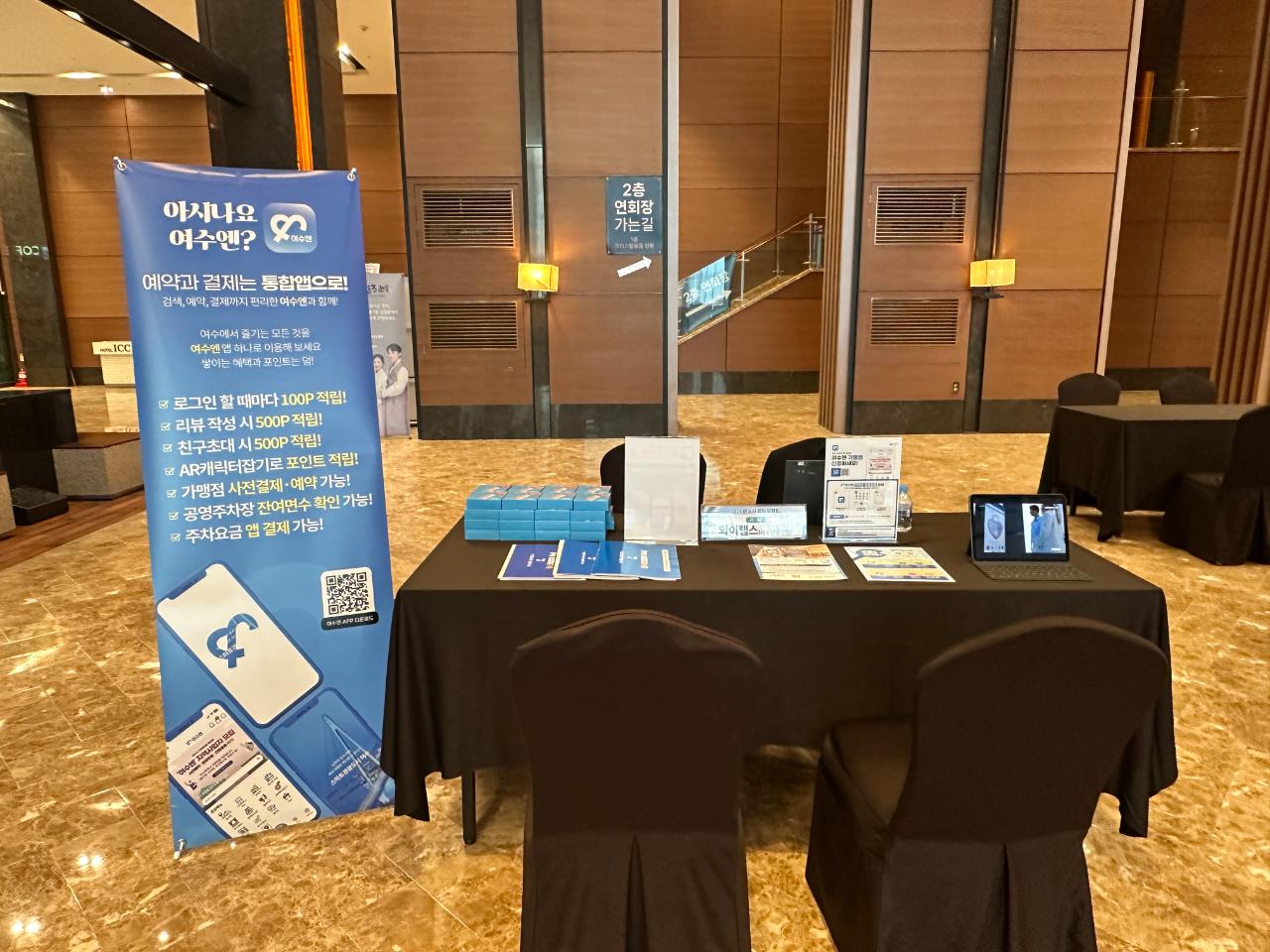 호텔 로비에 마련된 부스에서 어수 관련 예약 및 결제 서비스에 대한 안내가 담긴 현수막과 다양한 홍보 자료가 전시되어 있다.
