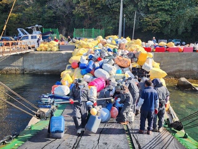 여수시 섬 지역에서 수거된 해양쓰레기를 작업선에 싣는 모습. 작업복을 입은 인력들이 스티로폼 부표, 플라스틱 통, 폐어구 등이 담긴 노란 자루와 부표류를 배 위로 옮기고 있다. 선착장에는 대량의 해양쓰레기 자루가 쌓여 있다.