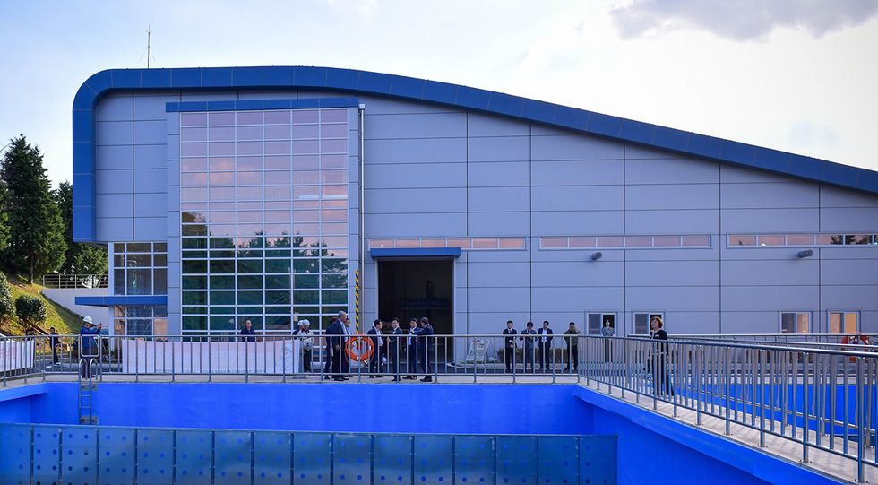 건물 앞에 여러 사람이 모여 있는 모습으로, 배경에는 큰 유리창이 있는 현대적인 건물이 보인다. 수영장처럼 보이는 푸른 물이 있는 공간이 있다.