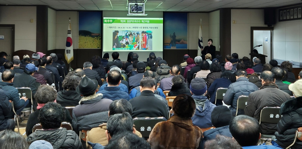 많은 사람들이 강당에서 발표를 듣고 있으며, 화면에는 제3회 산양유옥수수 팩스티벌에 대한 정보가 표시되고 있다.