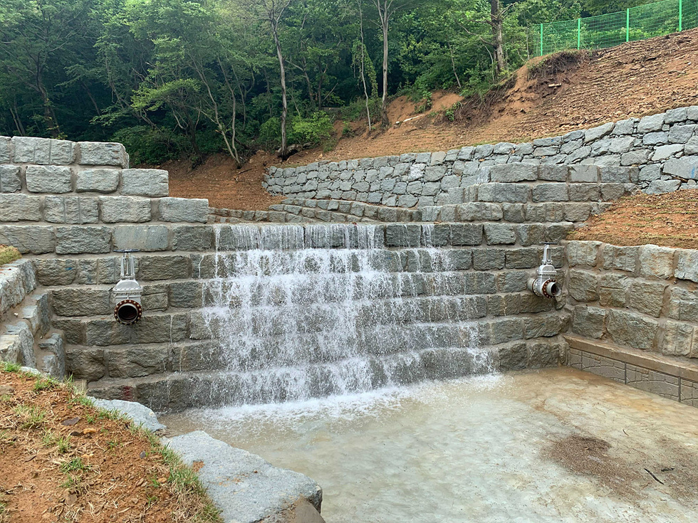 돌로 쌓인 구조물에서 물이 흐르는 장면으로, 주변은 나무와 흙으로 둘러싸여 있다.