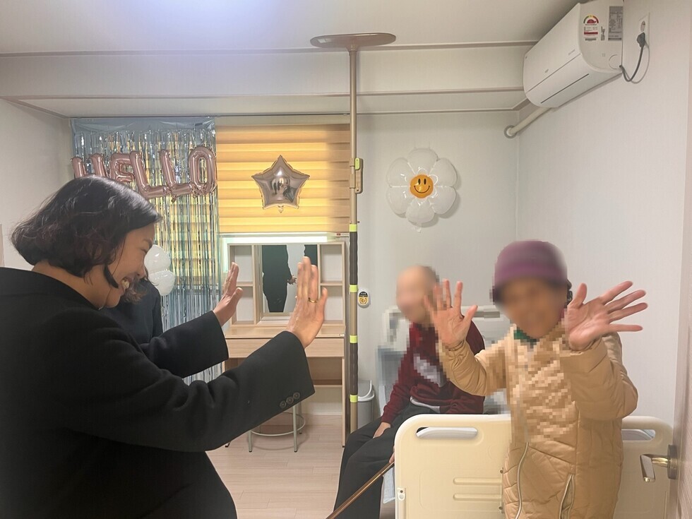 한 여성이 병원 침대 옆에 있는 두 사람과 함께 손을 흔들며 인사하고 있는 장면이다. 배경에는 헬로라는 글자가 적힌 풍선이 보인다.