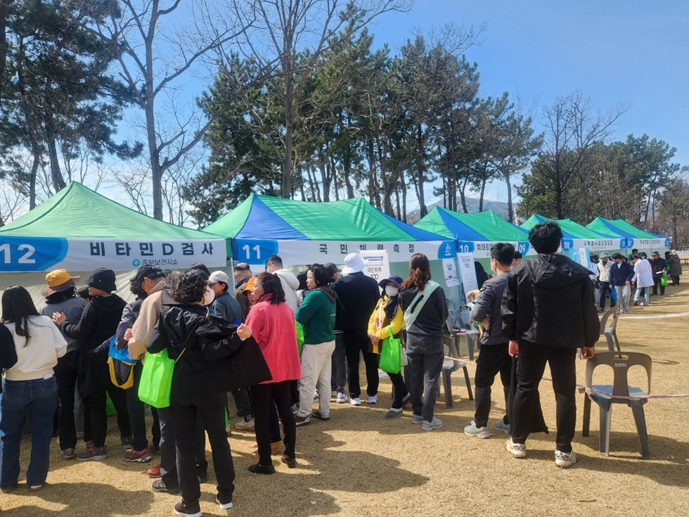사람들이 비타민 D 검사와 국민 혈압 측정을 받기 위해 대기하고 있는 장면으로, 여러 개의 텐트가 설치된 야외 행사 현장이다.