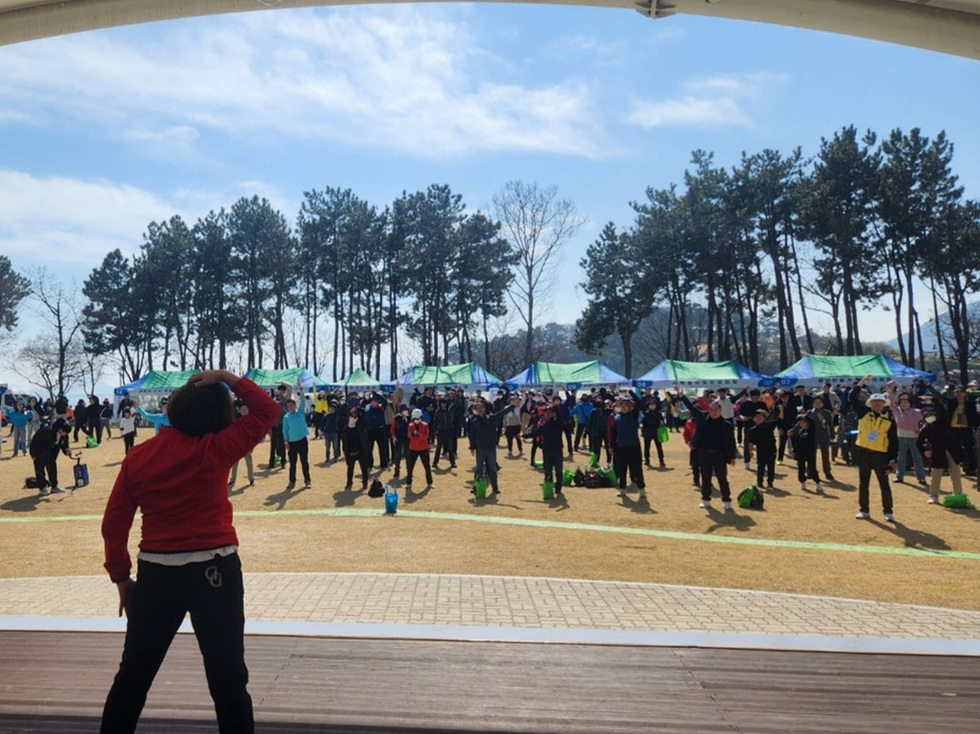 한 남성이 무대에서 관중을 향해 운동을 지도하고 있으며, 많은 사람들이 그를 따라 운동하고 있는 모습이다. 배경에는 나무와 텐트가 보인다.