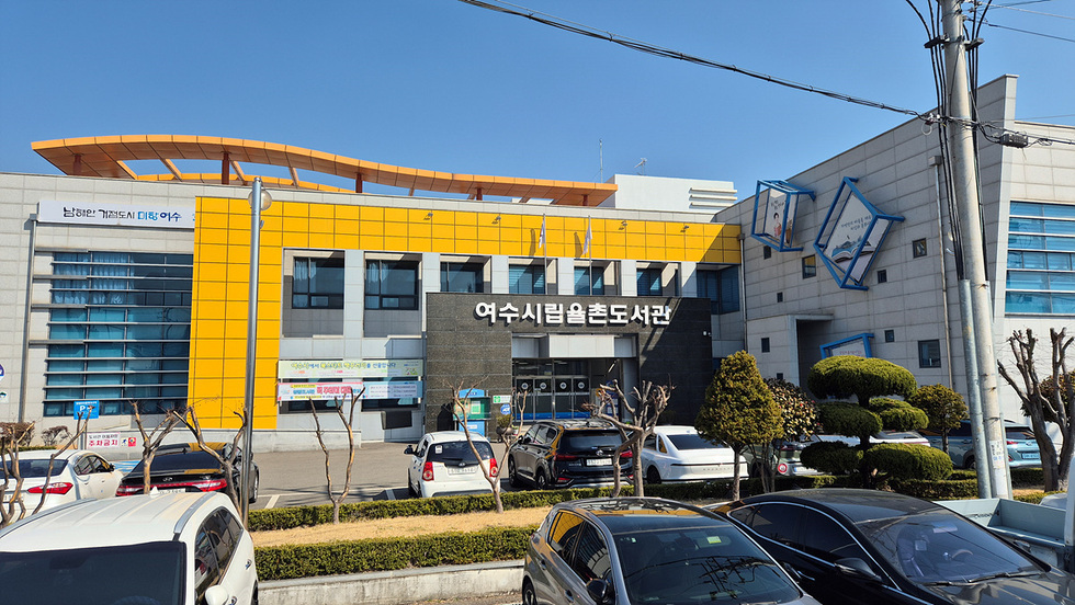 여수시립율촌도서관의 외관이 보이는 장면으로, 도서관 앞에는 주차된 차량들이 있으며, 맑은 하늘 아래 건물이 위치해 있다.