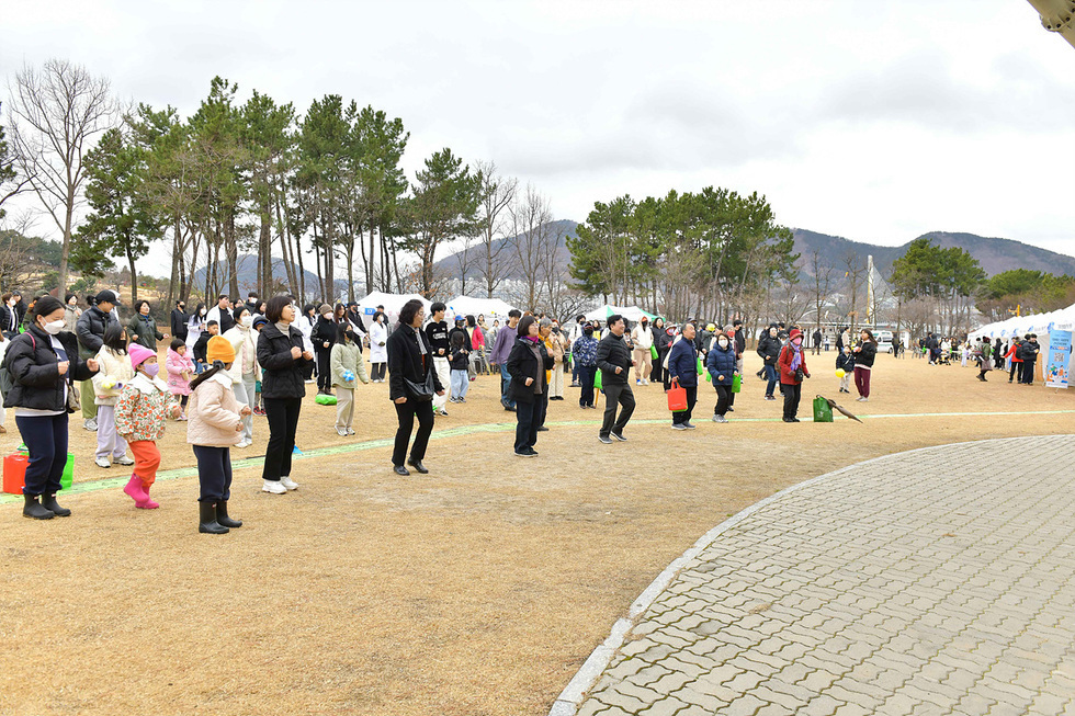 많은 사람들이 야외에서 운동을 하고 있으며, 어린이와 어른들이 함께 참여하고 있다. 배경에는 나무와 텐트가 보인다.