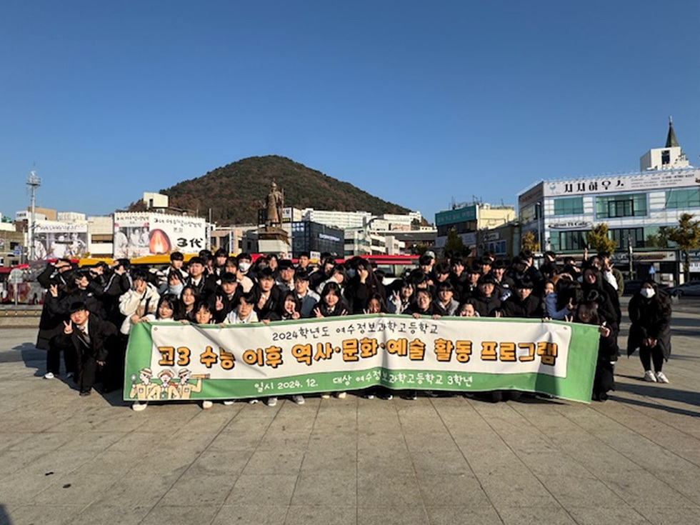 어울림 고등학교 학생들이 역사 문화 예술 활동 프로그램을 기념하며 단체 사진을 찍고 있다. 배경에는 동산과 상업 건물이 보인다.