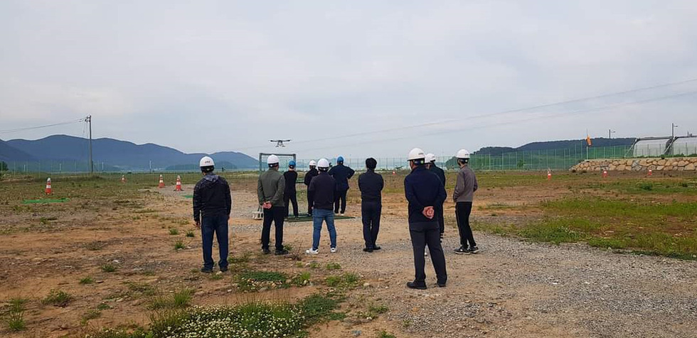 드론이 비행 중인 모습을 지켜보는 여러 명의 사람들이 야외에서 헬멧을 착용하고 서 있다. 배경에는 산과 울타리가 보인다.