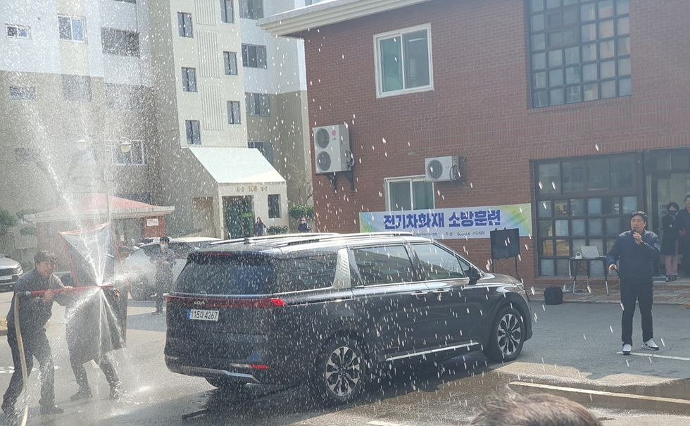 전기차 화재 소방 훈련이 진행되고 있는 모습으로, 한 남성이 차량 옆에서 설명하고 있으며, 다른 사람들이 소화기를 사용해 물을 뿌리고 있다.