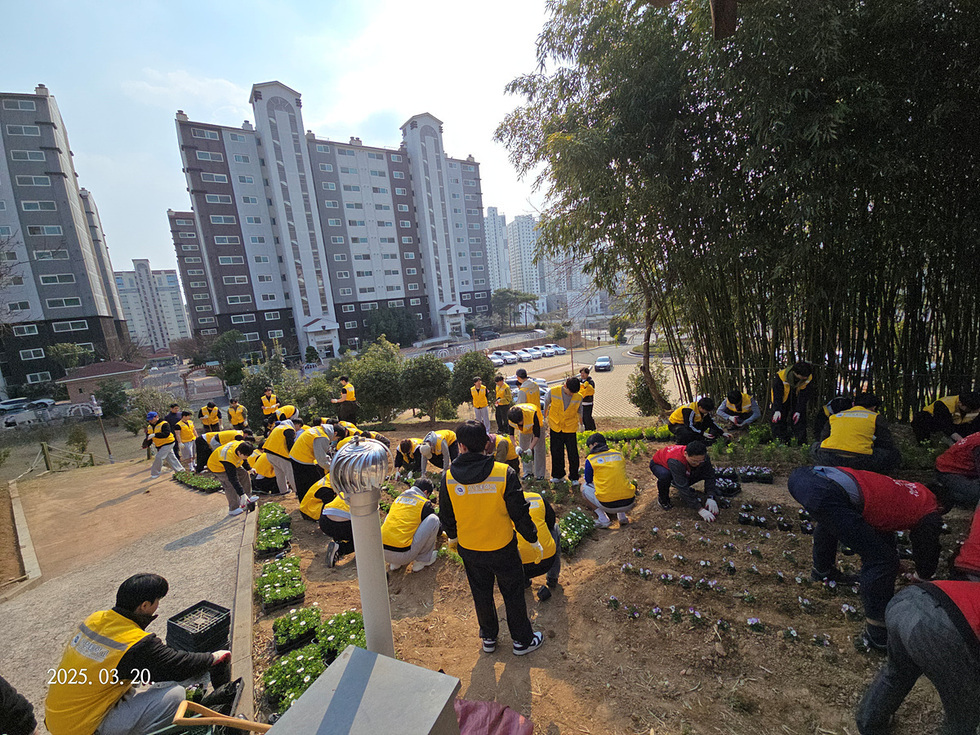 많은 사람들이 노란 조끼를 입고 꽃을 심고 있는 모습으로, 아파트 단지가 배경에 보인다.