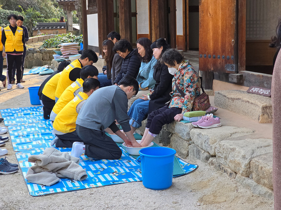 자원봉사자들이 노인들의 발을 씻겨주고 있는 장면으로, 노인들은 벤치에 앉아 있으며 자원봉사자들은 노란 조끼를 입고 있다.