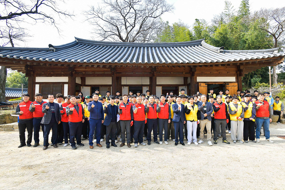 여러 사람이 전통 한옥 앞에서 단체 사진을 찍고 있으며, 일부는 손가락으로 하트를 만들고 있다. 이들은 자원봉사 활동에 참여한 것으로 보인다.