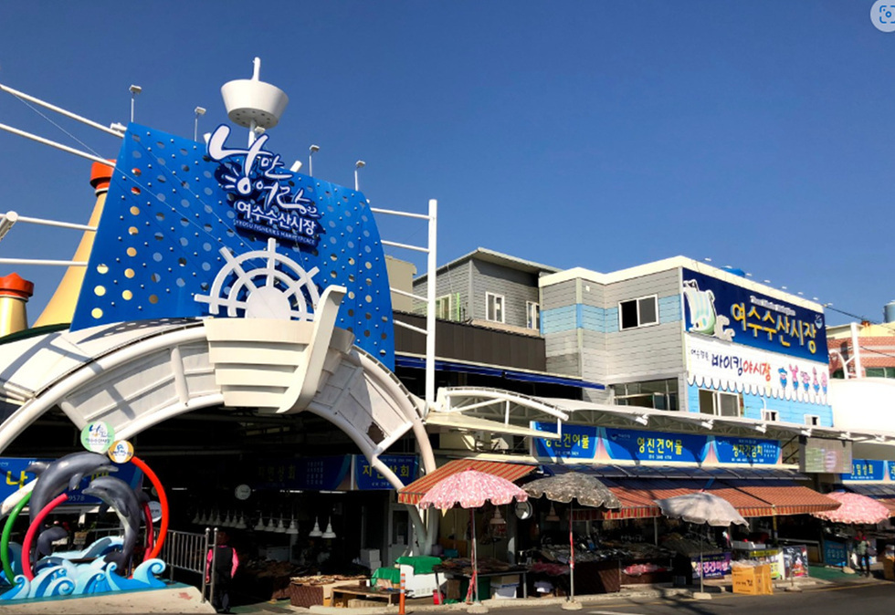 여수수산시장의 입구 모습으로, 파란색 간판에 여수수산시장이라는 글자가 적혀 있다. 주변에는 다양한 상점과 시장이 펼쳐져 있다.