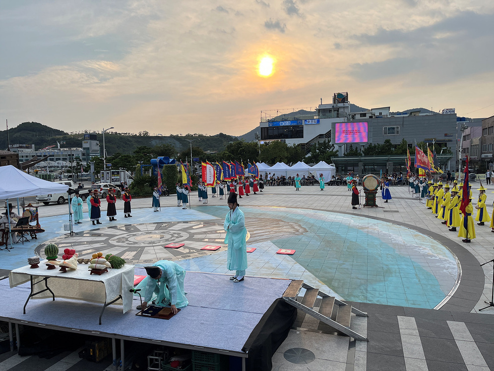 여러 사람이 전통 의상을 입고 공연을 준비하는 모습으로, 중앙에는 제물과 함께 무대가 마련되어 있다. 배경에는 해가 지고 있으며, 다양한 깃발이 세워져 있다.