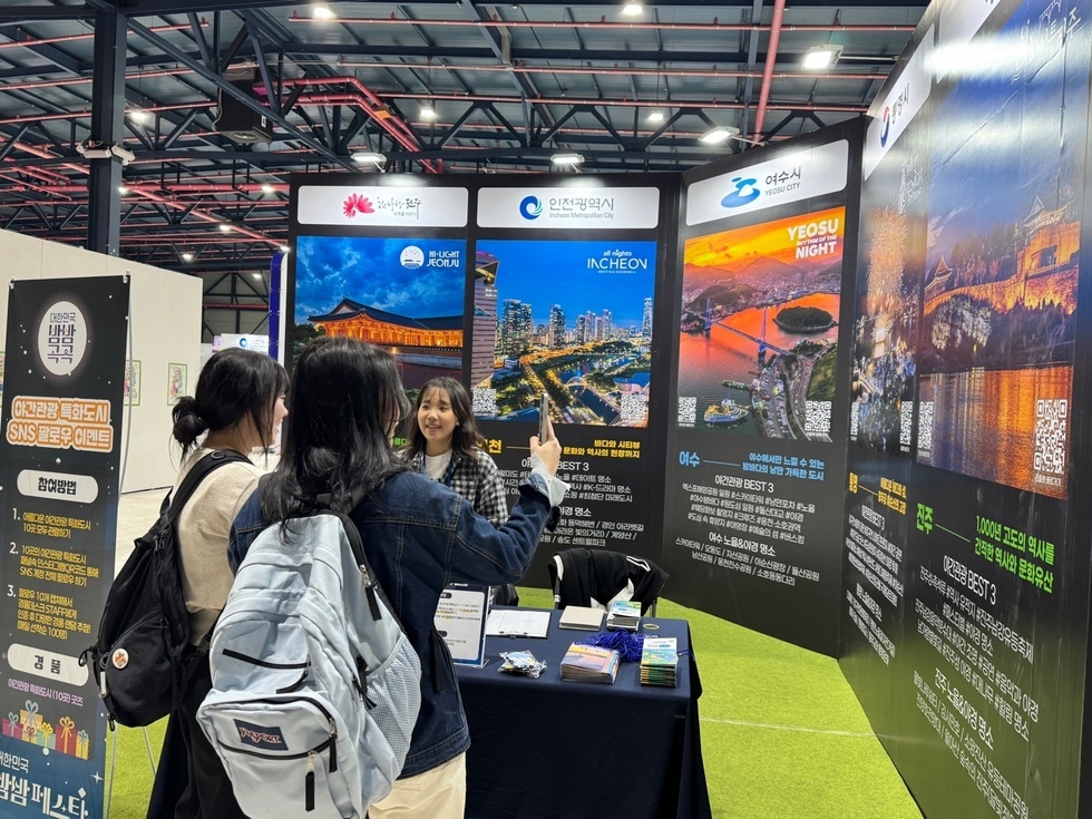 여행 박람회 부스에서 한 여성 직원이 방문객들과 대화하고 있으며, 부스에는 대한민국의 관광지와 관련된 정보가 담긴 배너가 세워져 있다.