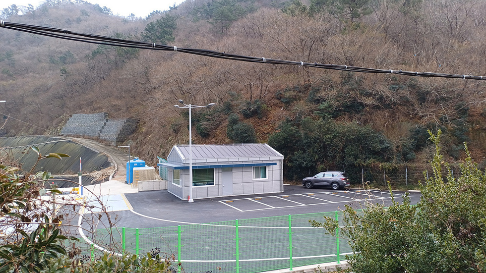 산기슭에 위치한 작은 건물과 주차장이 보인다. 주차장에는 차량이 한 대 주차되어 있으며, 주변은 나무와 덤불로 둘러싸여 있다.