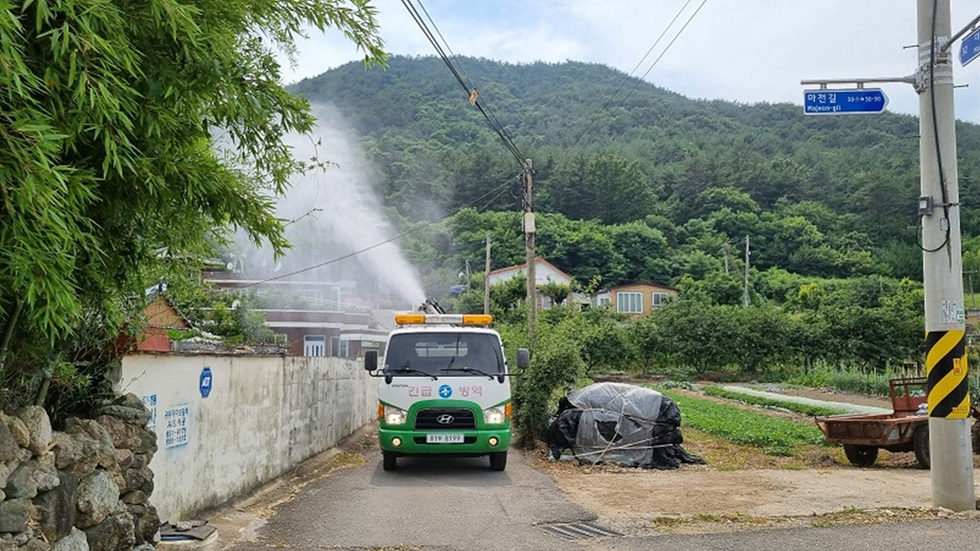 긴급 방역 차량이 마을 도로에서 방역 작업을 하고 있는 모습으로, 주변에는 나무와 집들이 보인다.