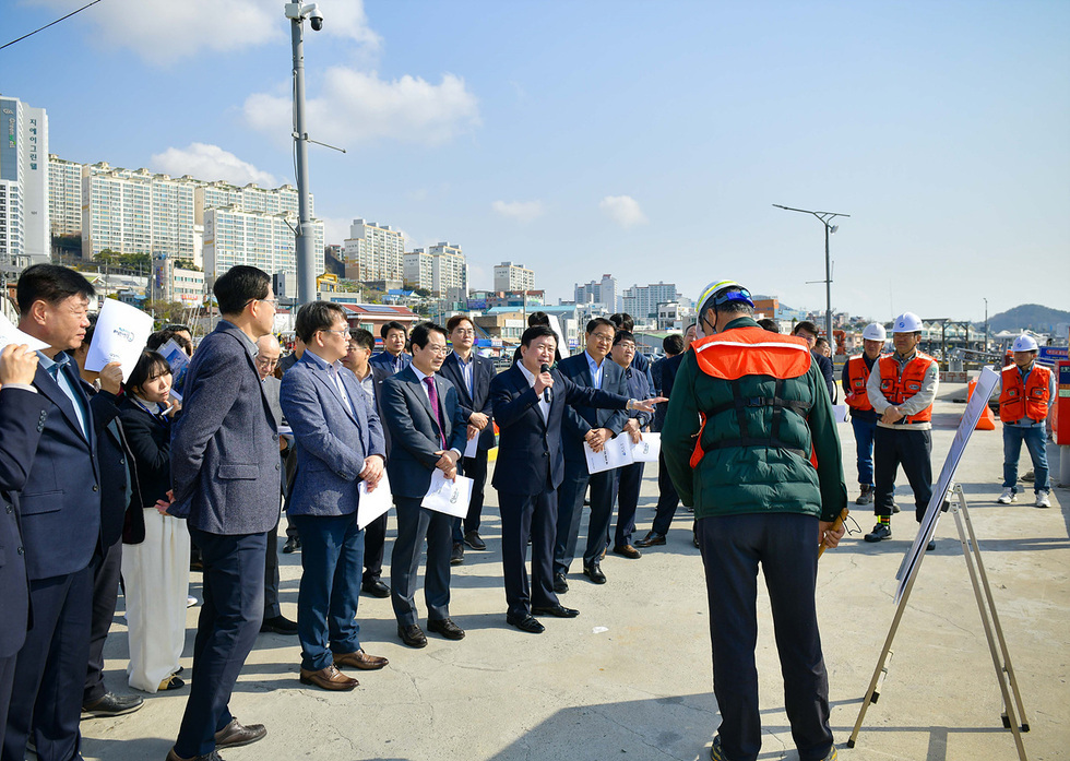 한 남성이 마이크를 들고 연설하고 있으며, 여러 명의 사람들이 그를 바라보고 있다. 배경에는 고층 건물과 항구가 보인다.