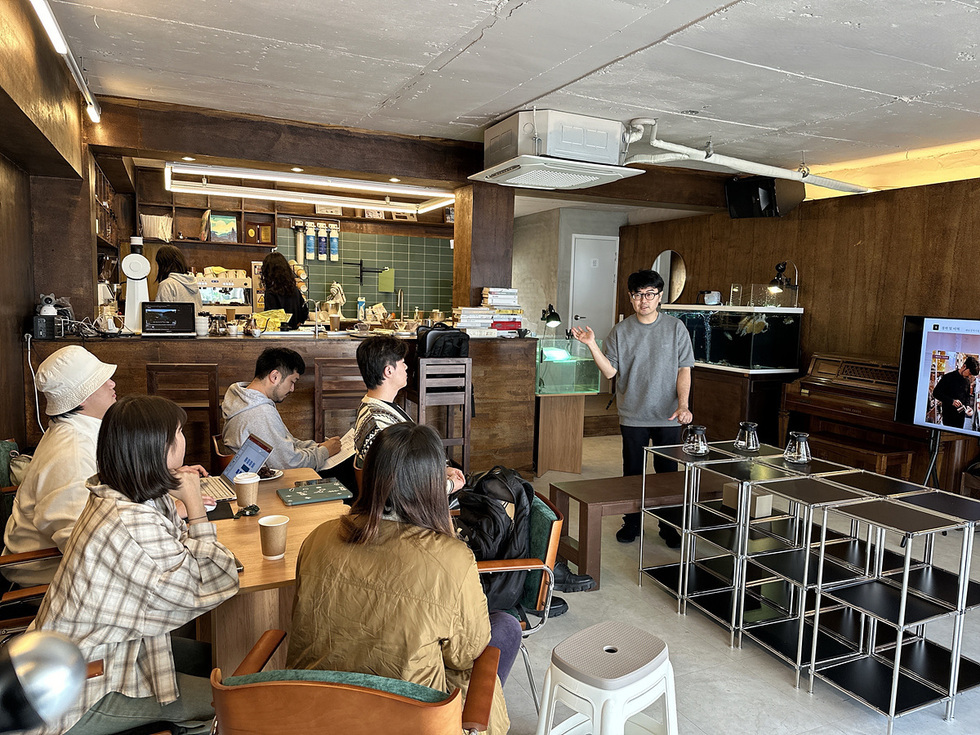 카페에서 한 남성이 청중에게 발표를 하고 있으며, 청중은 테이블에 앉아 그를 주의 깊게 듣고 있다. 배경에는 카페의 조리대와 여러 가지 음료가 보인다.