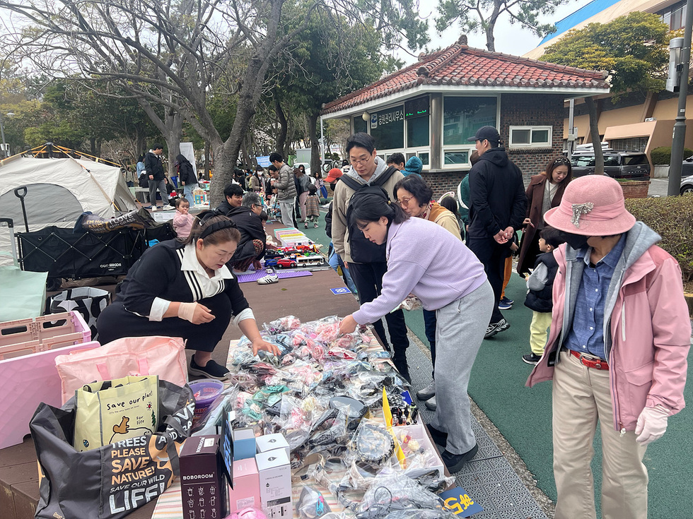 사람들이 야외에서 물건을 판매하고 있는 장면으로, 한 여성이 상품을 살펴보고 있다. 배경에는 공원 관리 사무소가 보인다.