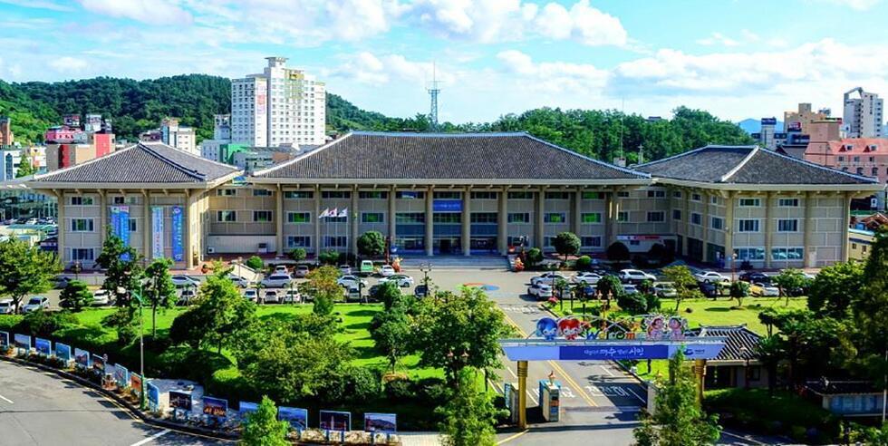 전통적인 건축 양식의 건물이 보이는 장면으로, 주변에 나무와 잔디가 있는 공원이 있다. 건물 앞에는 차량들이 주차되어 있으며, 배경에는 도시의 고층 건물들이 보인다.