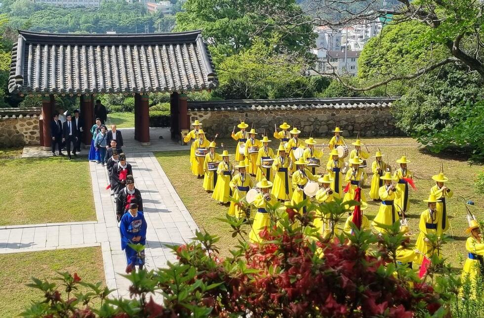전통 의상을 입은 사람들이 노란색 옷을 입고 행진하며 공연을 준비하고 있는 모습이다. 배경에는 고풍스러운 건물과 푸른 나무들이 보인다.