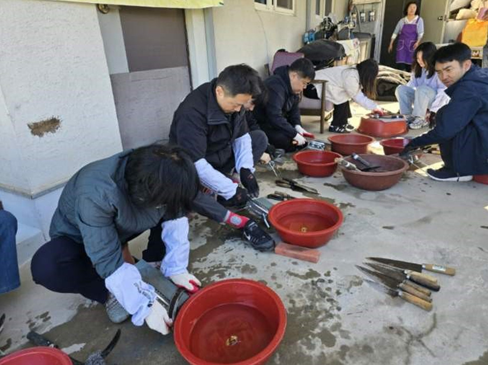 여러 사람이 빨간 그릇에 무엇인가를 준비하며 작업하고 있는 모습이다. 배경에는 다른 사람들이 보이고, 작업 도구들이 바닥에 놓여 있다.