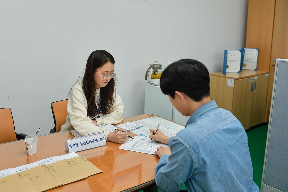 여성 직원이 남성 고객과 상담하며 서류를 작성하는 장면으로, 테이블 위에는 여러 문서와 필기도구가 놓여 있다.