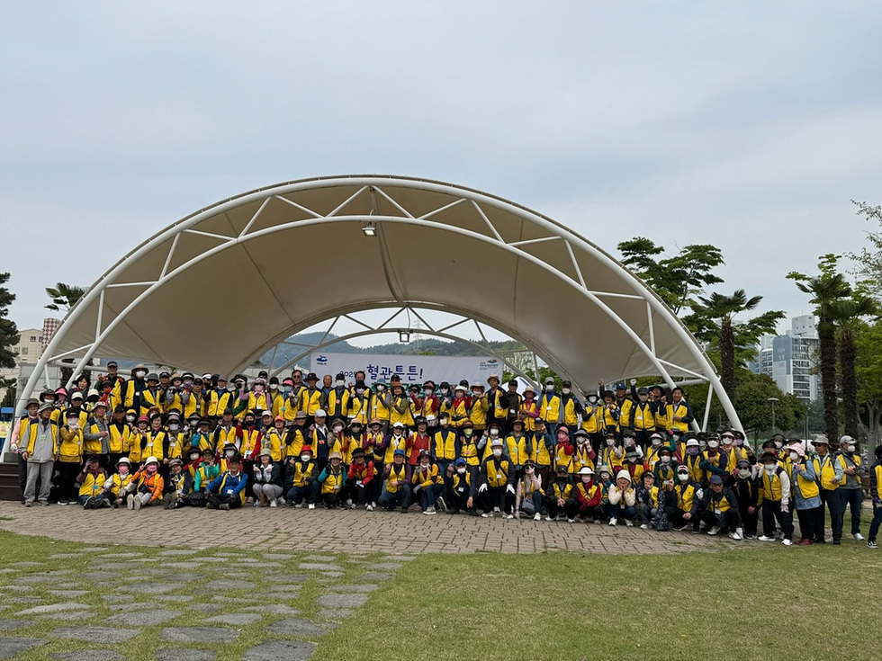 많은 사람들이 노란 조끼를 입고 야외에서 단체 사진을 찍고 있다. 배경에는 형관트 행사 관련 현수막이 보인다.