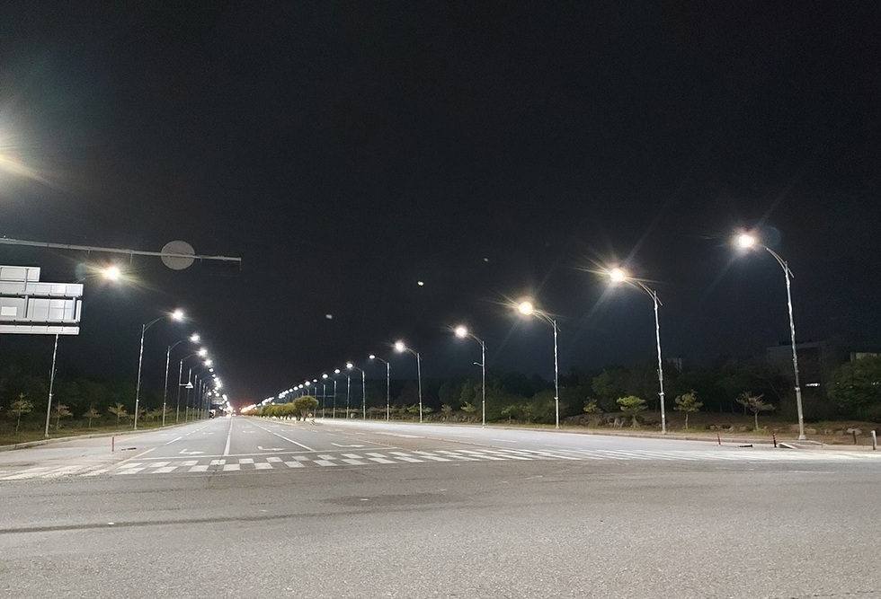 어두운 밤에 가로등이 줄지어 서 있는 도로의 모습이다. 도로는 한적하며, 멀리 신호등과 도로 표지판이 보인다.