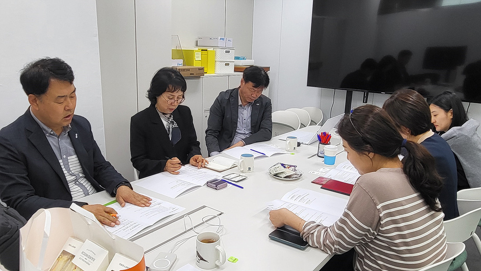 회의 중인 여러 명의 사람들이 테이블에 앉아 문서에 집중하고 있다. 배경에는 모니터와 사무용품이 보인다.