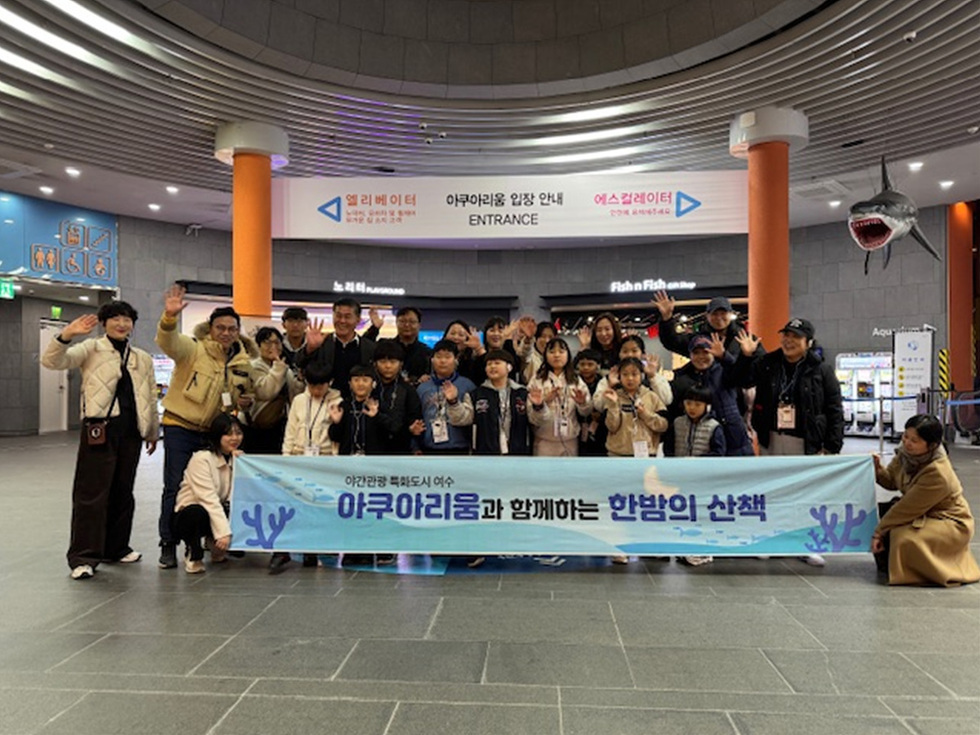 아쿠아리움에서 한밤의 산책 프로그램에 참여하는 어린이들과 어른들이 함께 사진을 찍고 있다. 배경에는 아쿠아리움 입구와 관련된 안내 표지판이 보인다.