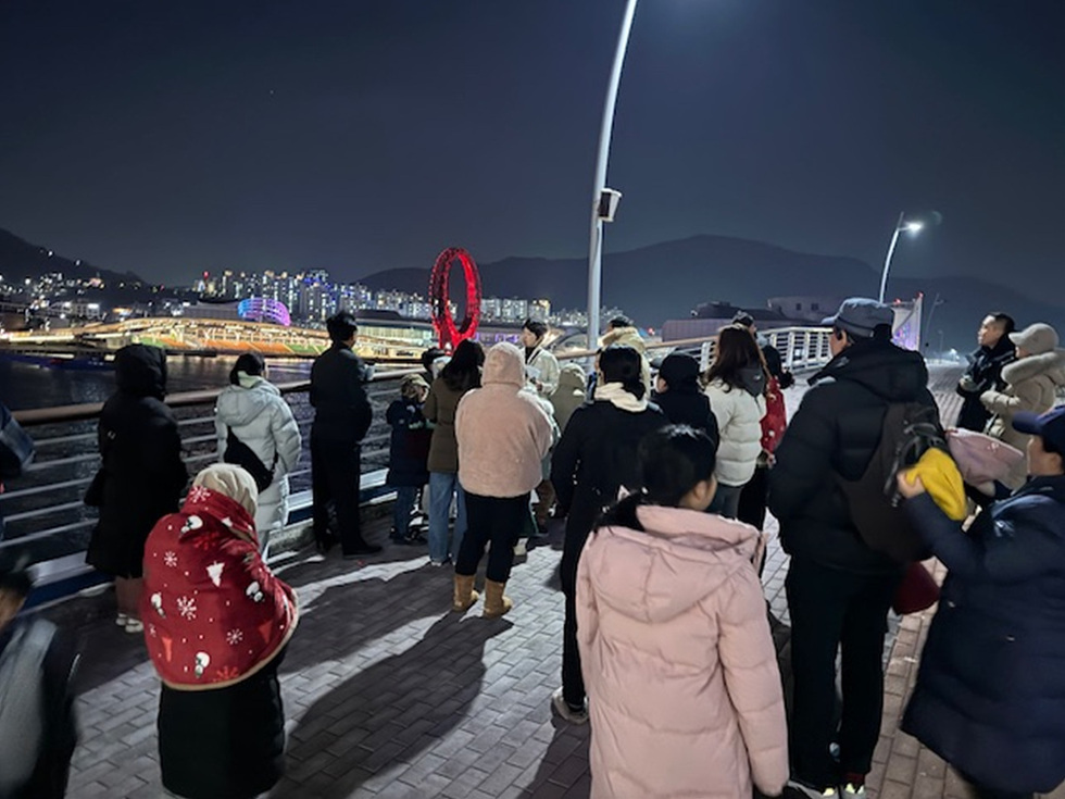 밤에 사람들이 다리 위에서 경치를 감상하고 있으며, 배경에는 조명이 켜진 도시와 대관람차가 보인다.