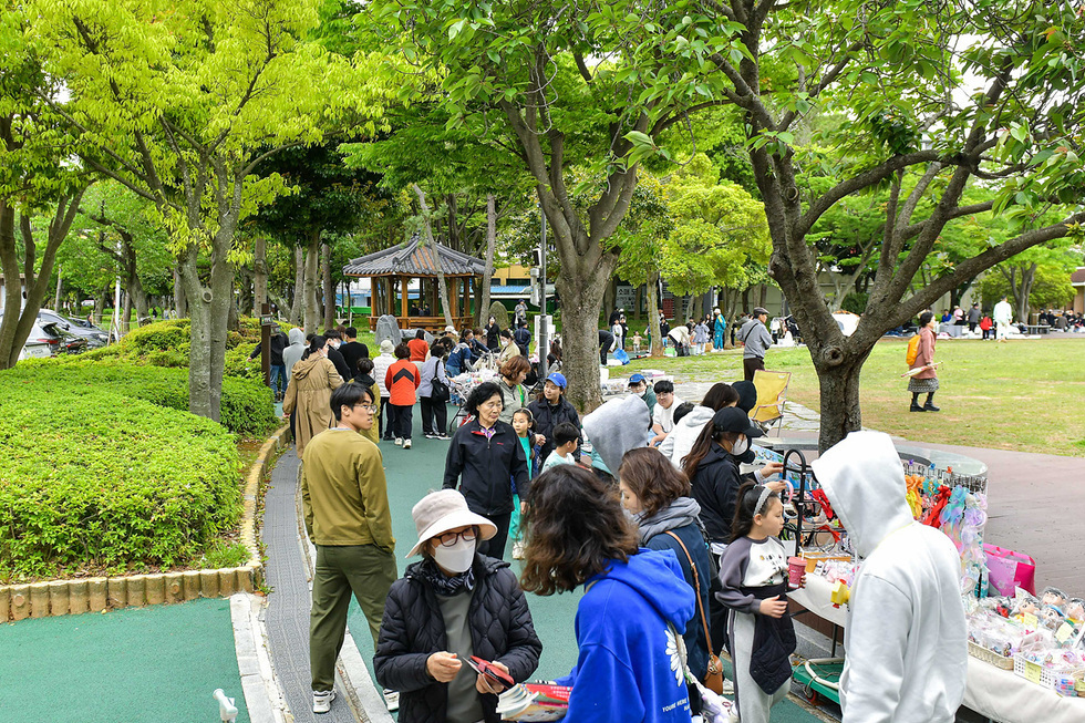사람들이 공원에서 모여 다양한 물건을 사고 있는 모습이다. 중앙에는 정자가 보이고, 주변에는 나무와 풀이 있다.