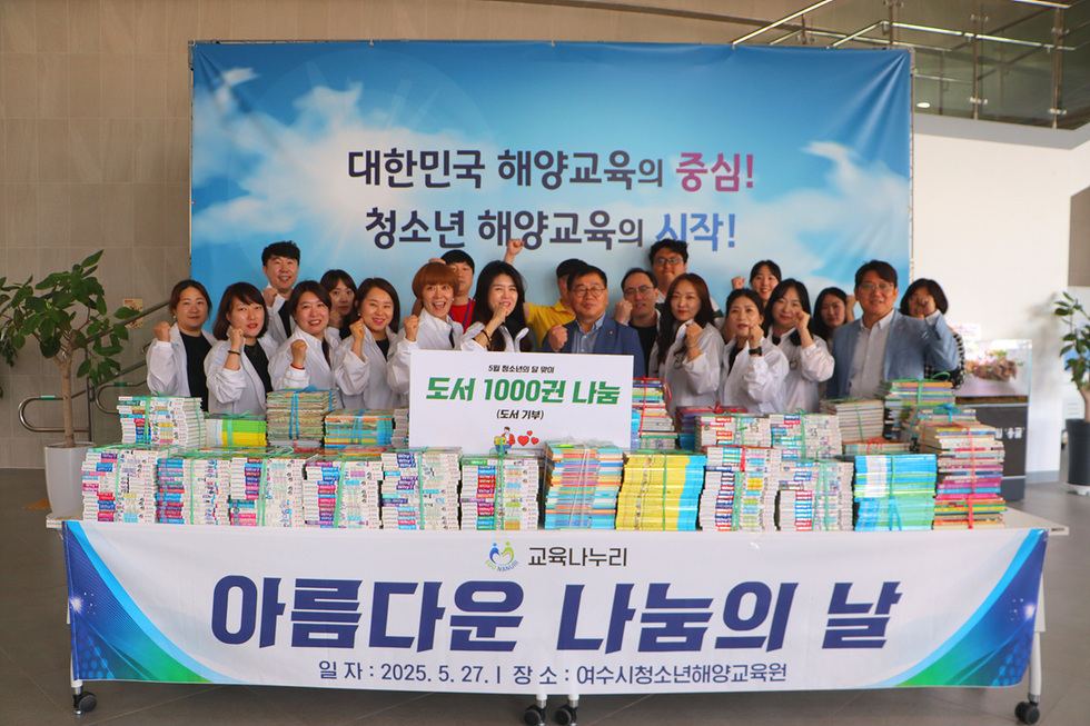 여수시청소년해양교육원에서 도서 기부 행사에 참여한 사람들과 함께 기념사진을 찍고 있다. 배경에는 해양교육 관련 슬로건과 도서 기부에 대한 정보가 적힌 현수막이 있다.