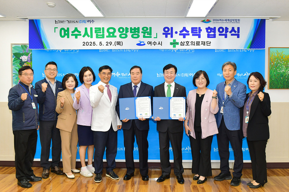 여수시립요양병원 위수탁 협약식에서 관계자들이 기념사진을 찍고 있다. 참석자들은 협약서 두 개를 들고 있으며, 모두 힘찬 주먹을 쥐고 있다.