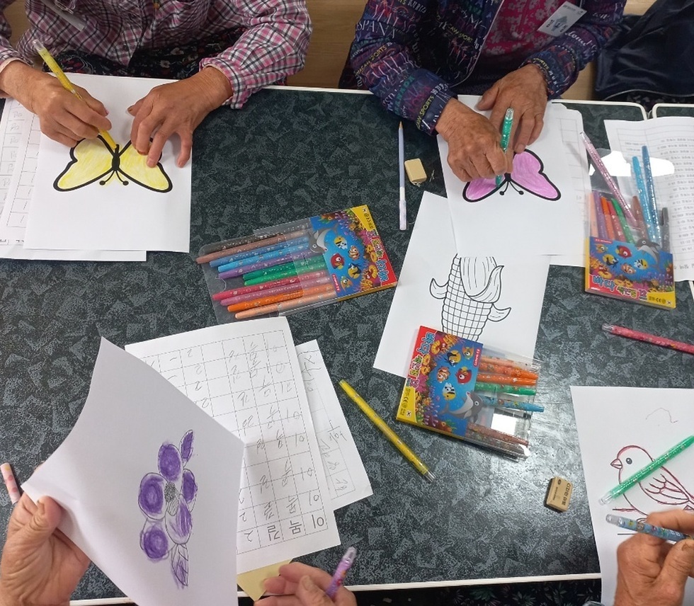 어르신들이 테이블에 앉아 색연필로 그림을 그리고 있는 모습으로, 다양한 색상의 색연필과 그림이 놓여 있다.