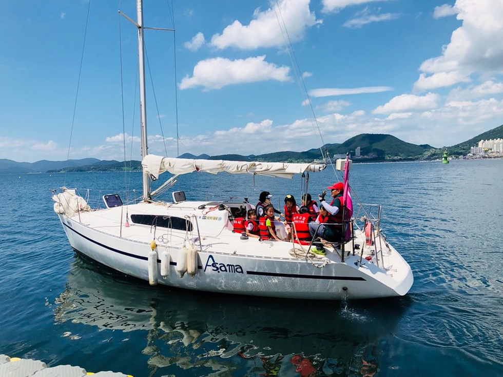 요트 Asama에서 구명조끼를 입은 사람들이 바다 위에서 즐거운 시간을 보내고 있다. 배경에는 푸른 산과 하늘이 펼쳐져 있다.