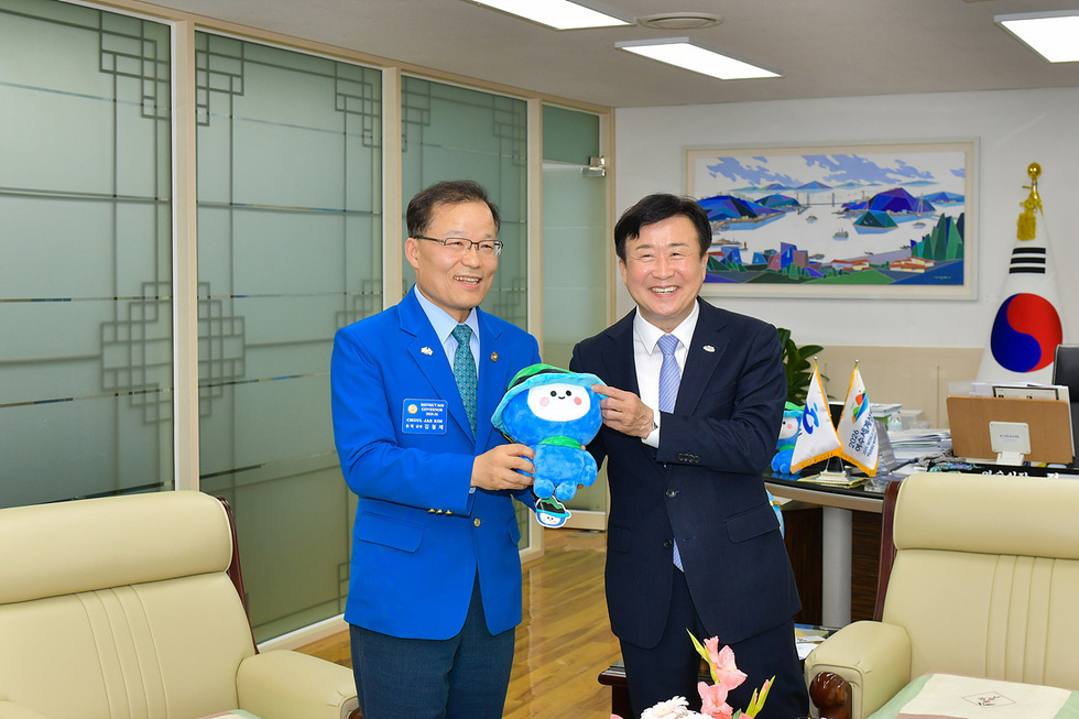 두 남성이 사무실에서 서로 미소를 지으며 인형을 교환하는 장면이다. 한 남성은 파란색 재킷을 입고 있으며, 다른 남성은 정장 차림이다.