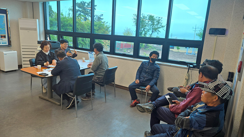 사람들이 회의 중인 실내 장면으로, 테이블에 앉아 있는 사람들과 벽에 기대어 있는 사람들이 있다. 창 밖으로 나무와 하늘이 보인다.