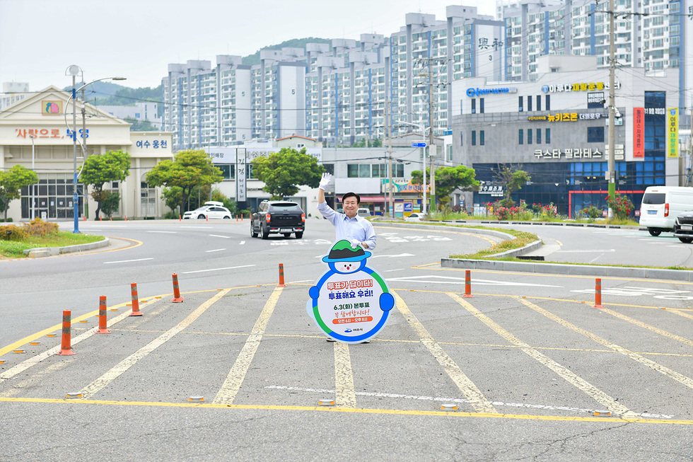 한 남성이 도로 중앙에 서서 투표를 독려하는 모습으로, 배경에는 아파트와 상점들이 보인다.
