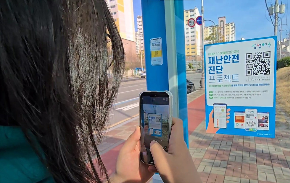 한 여성이 재난안전 진단 프로젝트 안내문을 스마트폰으로 촬영하고 있는 모습이다. 안내문에는 QR코드와 함께 비대면 안전교육에 대한 정보가 담겨 있다.